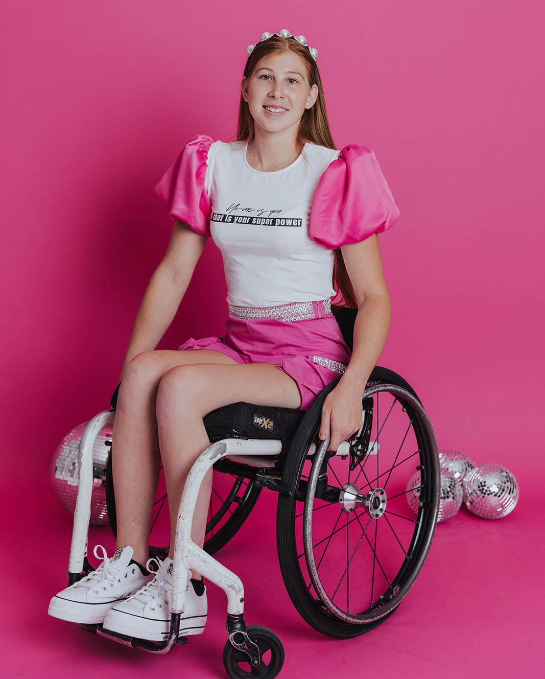 Young white women in a wheelchair posing in front of a pink background with some disco balls on the floor. She has red hair pulled back with a pearl headband. She is wearing a white tee with an inspirational quote on it ("No-One is You & That's Your Superpower") and bright pink princess puff sleeves. Her shorts are high-waisted pink cargo shorts with silver embellishments along the high waistline and pockets.