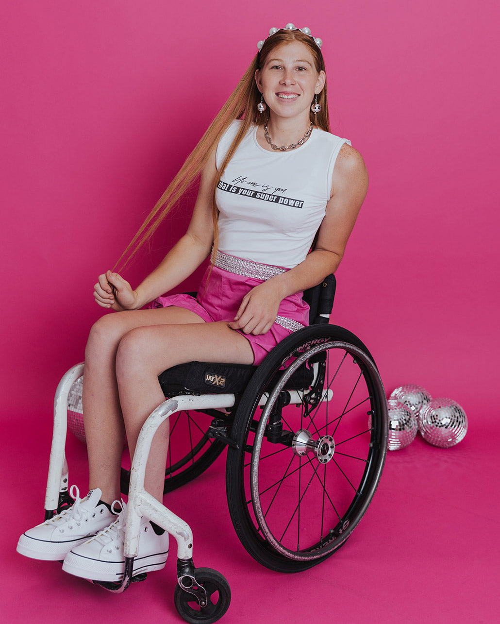 Young white woman with long red hair poses in a wheelchair in front of a pink background. She's holding out a strand of her hair while wearing a pearl headband. She is wearing a sleeveless white shirt, pink cargo shorts with silver embellishments and a high waist, and white sneakers.